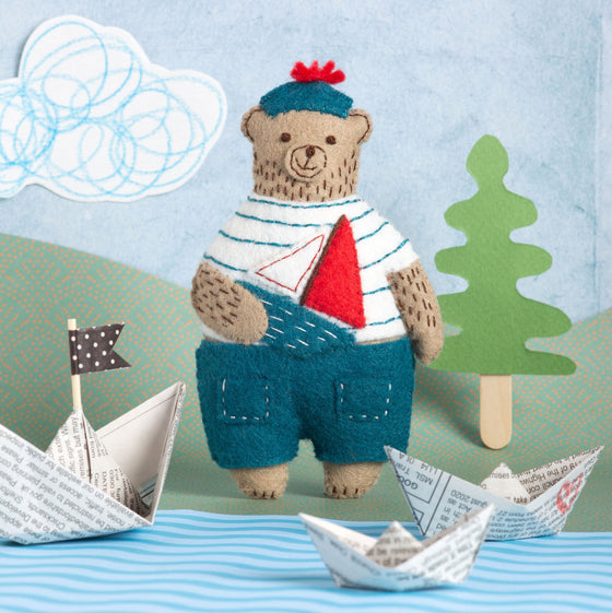Corinne Lapierre Felt Craft Mini Kit. A bear is stood, wearing a white t shirt with blue stripes.dark blue trousers and hat. The hat has a red pom pom. He is holding a toy sailing boat.