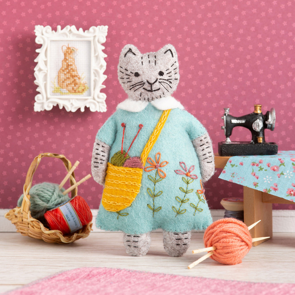 Corinne Lapierre Felt Craft Mini Kit. A grey cat with some black stitching on face, arms and legs, is stood. The cat is wearing a light turquoise dress and a yellow shoulder bag.